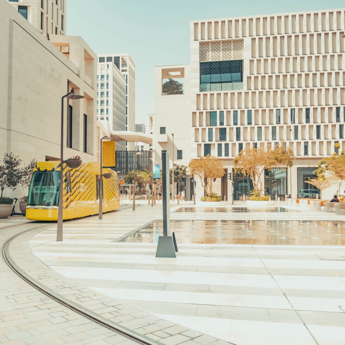 A modern yellow tram navigates tracks through a contemporary, sunlit urban plaza featuring geometric limestone buildings and a central water fountain.