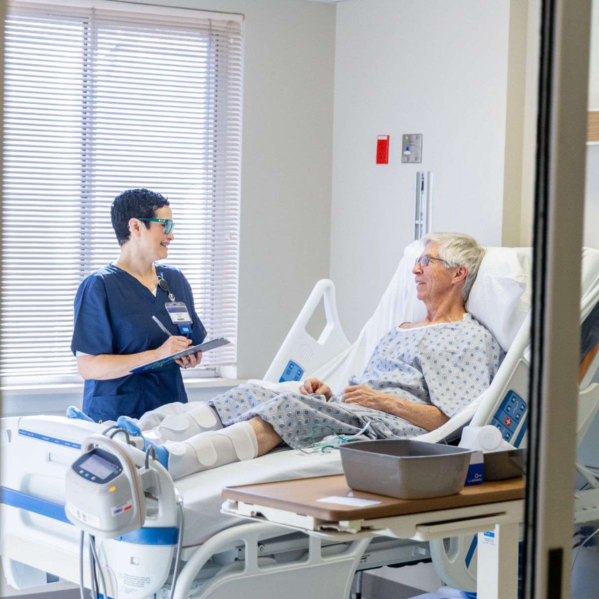 A friendly nurse in scrubs consults with an elderly patient resting in a hospital bed, focusing on compassionate healthcare communication.