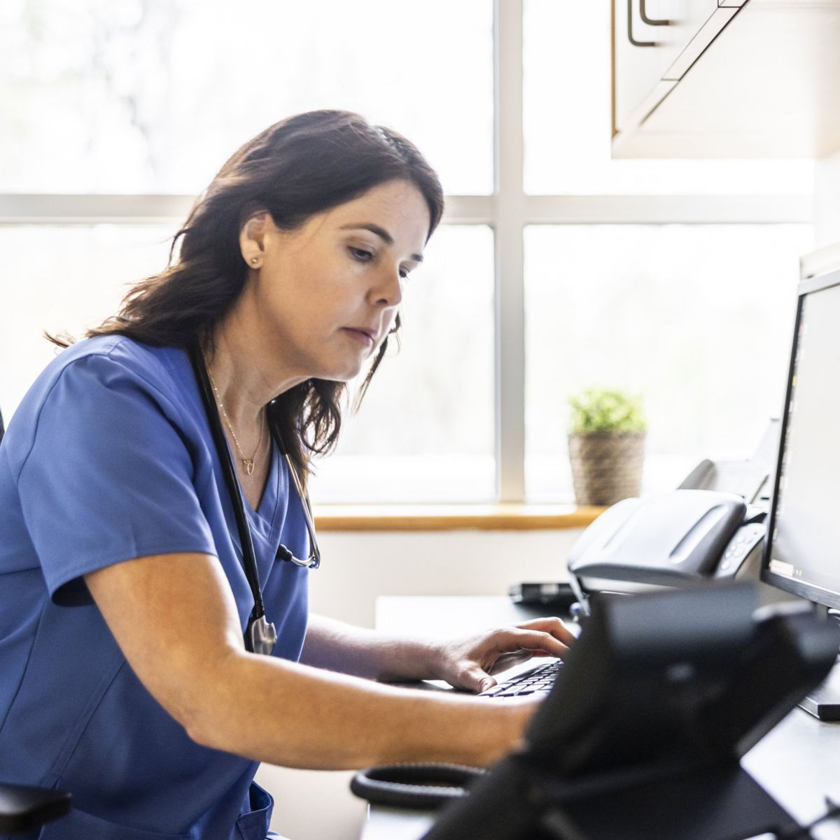 A focused nurse in blue scrubs types at a computer station in a bright, modern medical office setting.