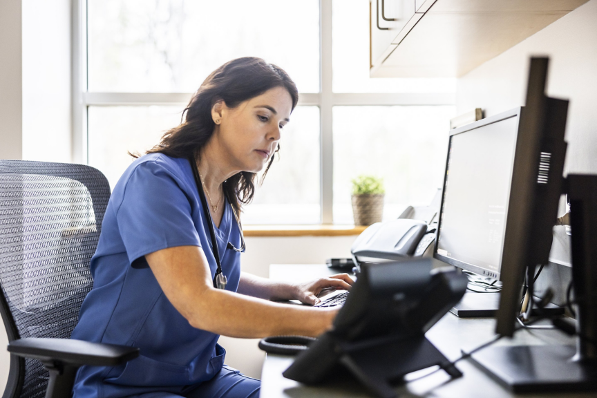 A focused nurse in blue scrubs types at a computer station in a bright, modern medical office setting.