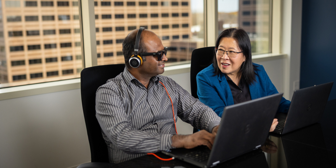 Two professionals working on laptops in a meeting room with large windows and office buildings visible in the background.