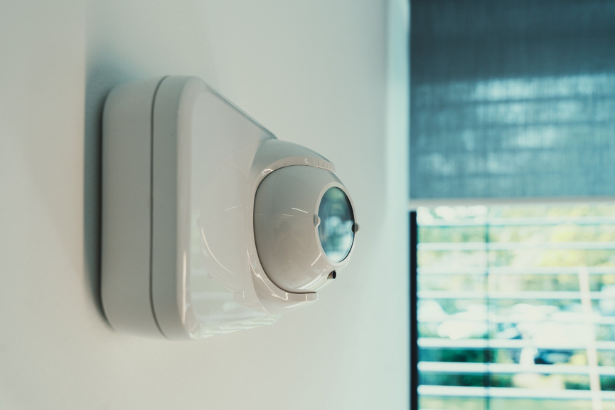 A close-up side profile of a modern white motion detection sensor mounted on a clean office wall near a window.