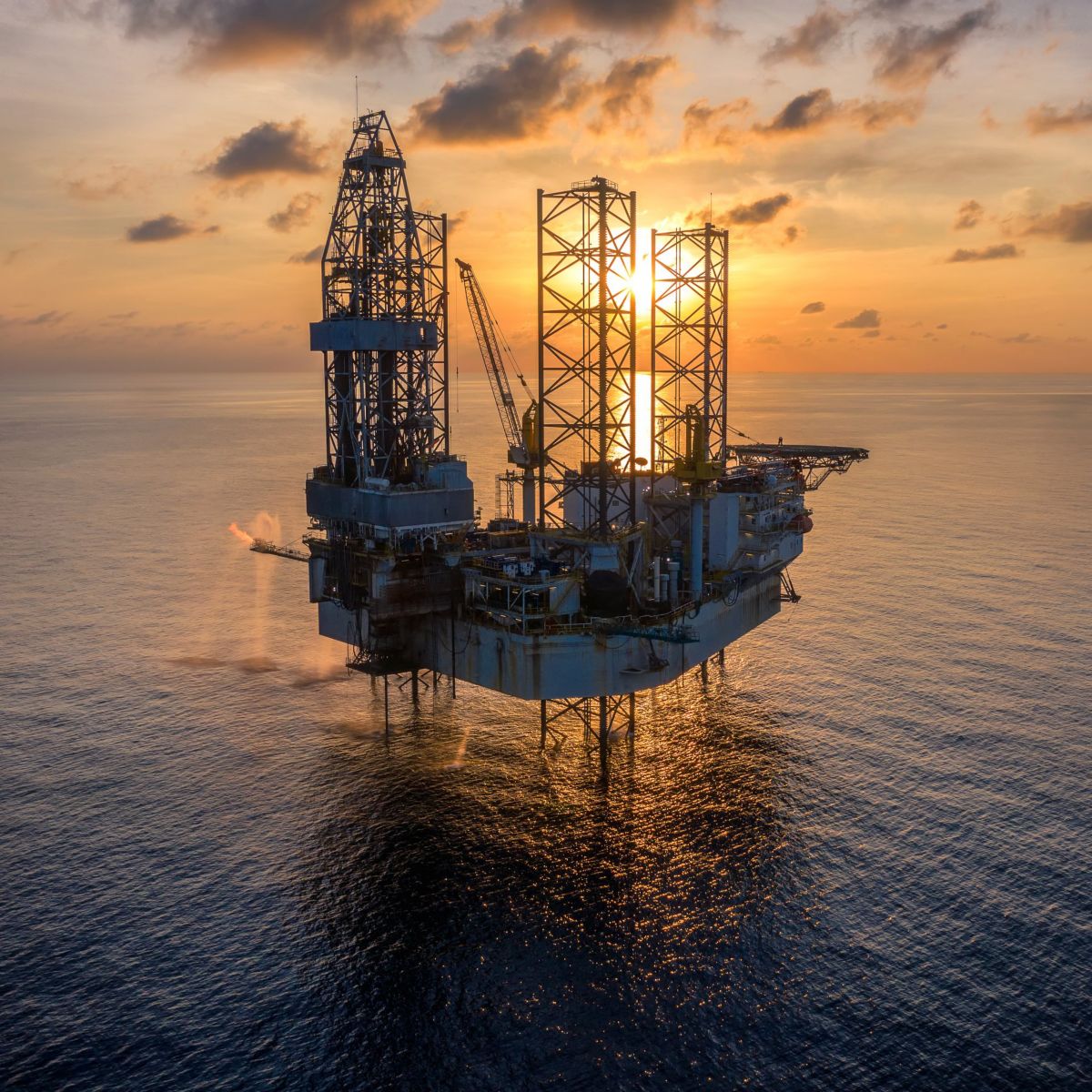 An aerial view of an offshore jack-up drilling rig silhouetted against a golden sunrise over the calm ocean.