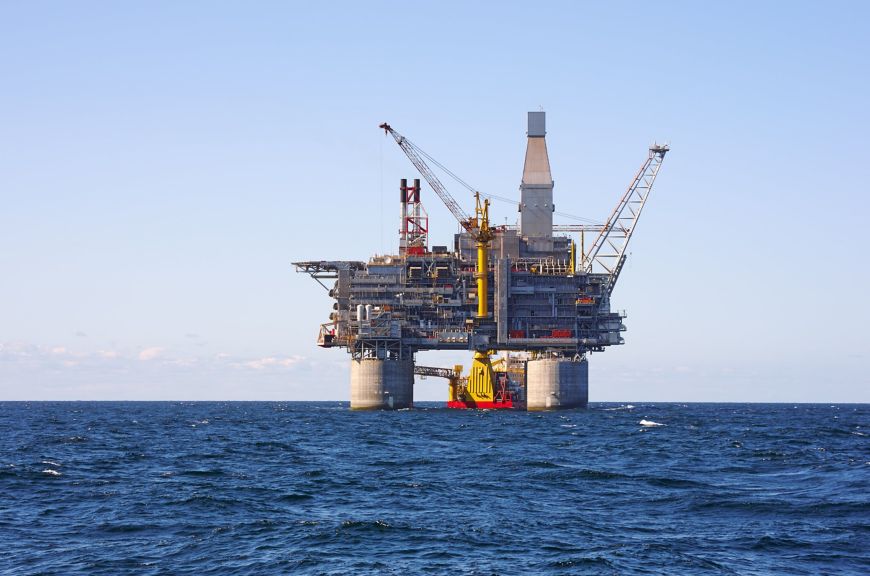 An industrial oil production platform stands in the middle of a vast, deep blue ocean under a clear sky.