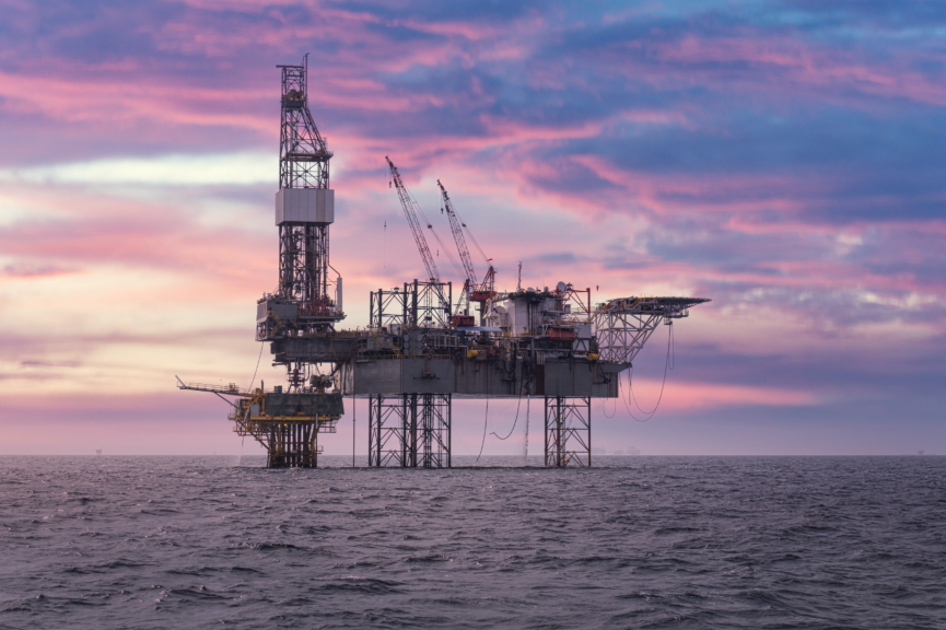 Ocean drilling platform on metal legs with cranes and equipment under a colorful sunset sky.