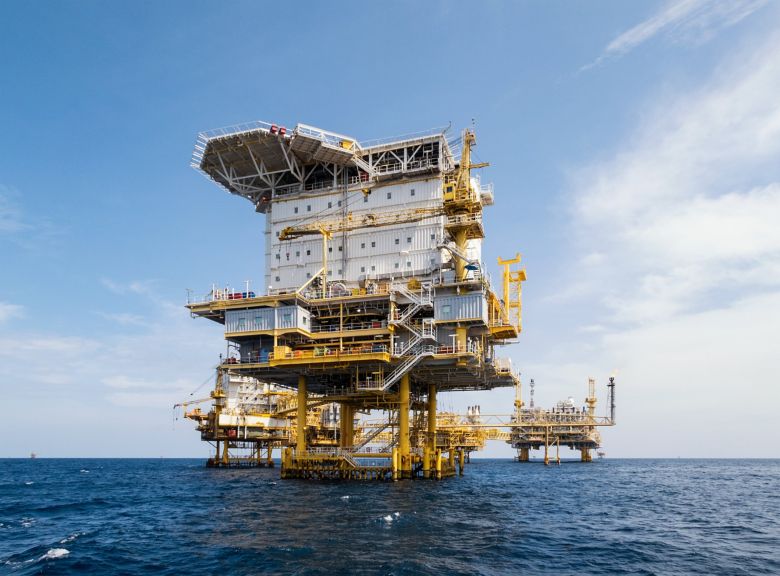 A large industrial offshore oil and gas production platform stands prominently in the middle of the deep blue ocean under a clear sky.
