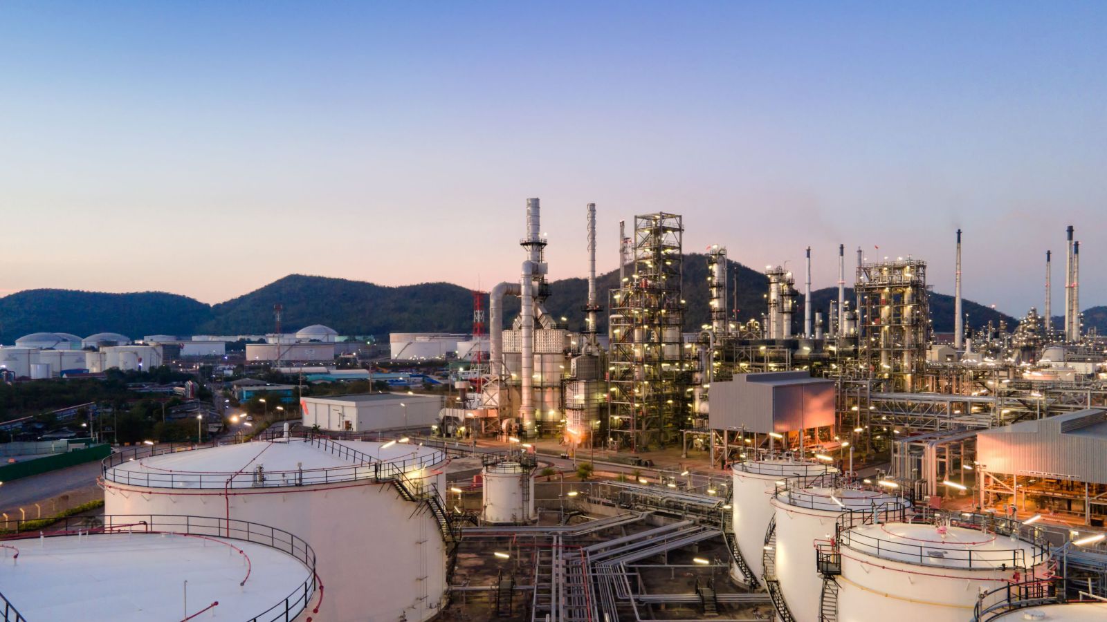 Beautiful sunset view of an oil refinery with illuminated structures and surrounding mountains at dusk.
