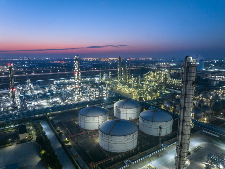 Aerial view of a petrochemical industrial plant illuminated against a twilight sky featuring large storage tanks and processing towers.
