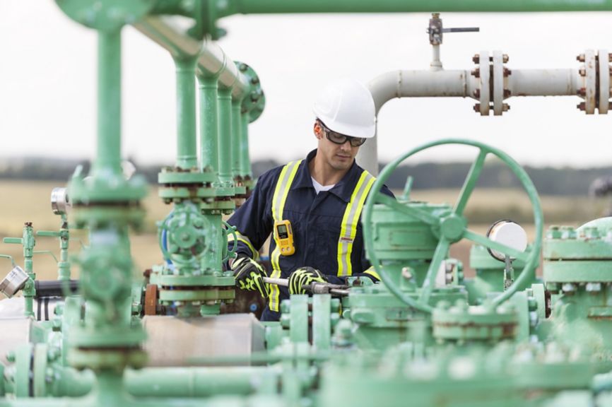 An industrial worker in safety gear uses a pipe wrench to perform maintenance on green infrastructure at an oil and gas field.