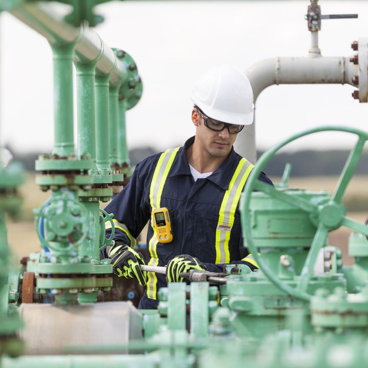 An industrial worker in safety gear uses a pipe wrench to perform maintenance on green infrastructure at an oil and gas field.
