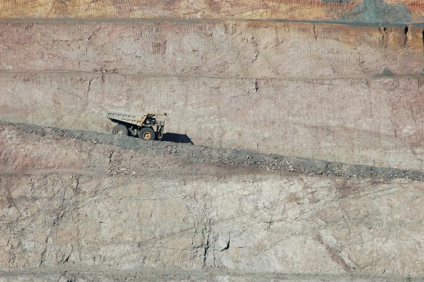 A heavy-duty haul truck traverses a diagonal path across the terraced rock walls of a large-scale open-pit mining operation.