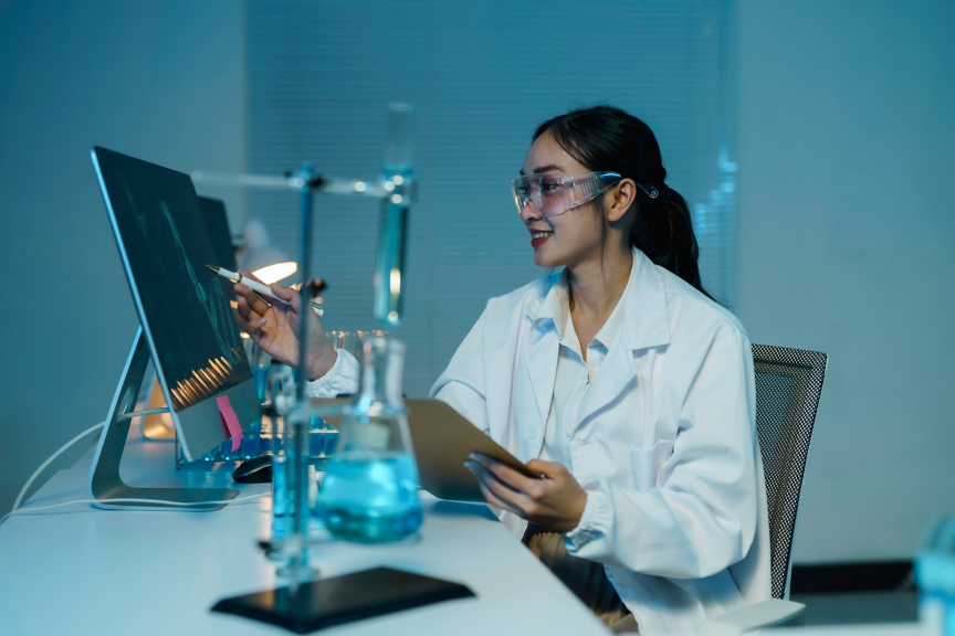 Smiling scientist wearing lab coat and protective glasses pointing at computer screen with pen and holding clipboard in modern laboratory with test tubes and beakers