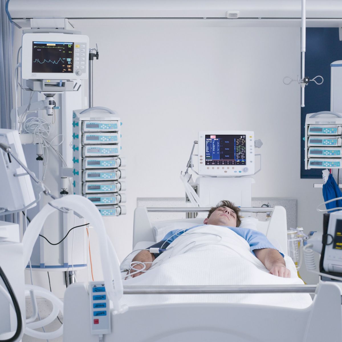 A man rests in a hospital bed surrounded by advanced life-support equipment and patient monitoring displays in a high-tech intensive care unit.