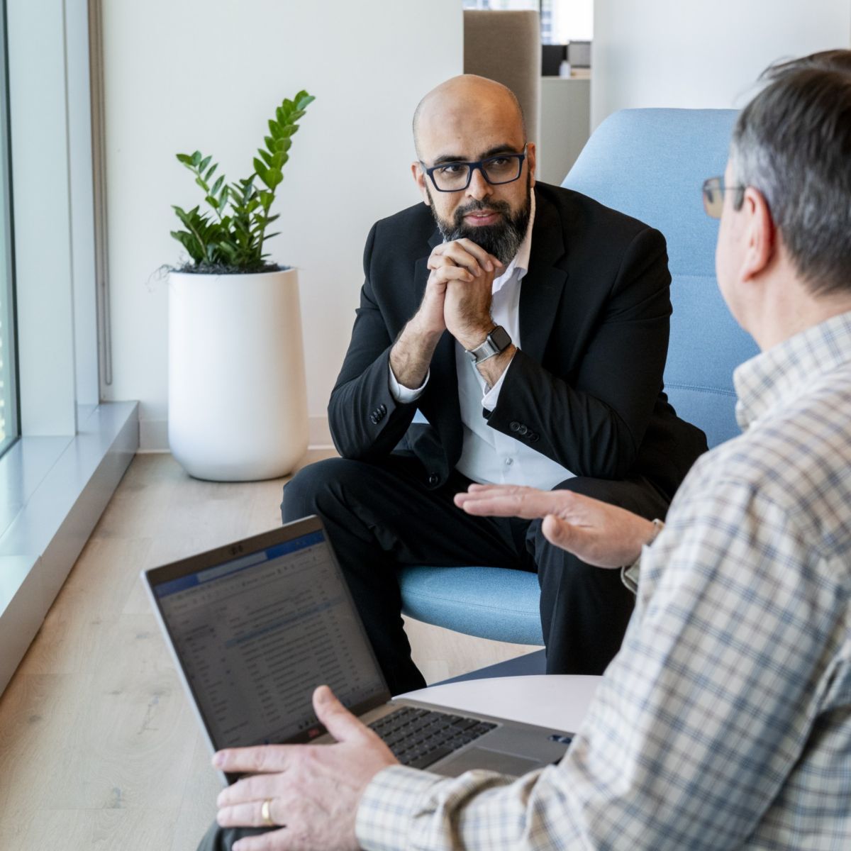 Two people seated facing each other, one using a laptop and the other listening, engaged in a discussion near a window in an office setting.