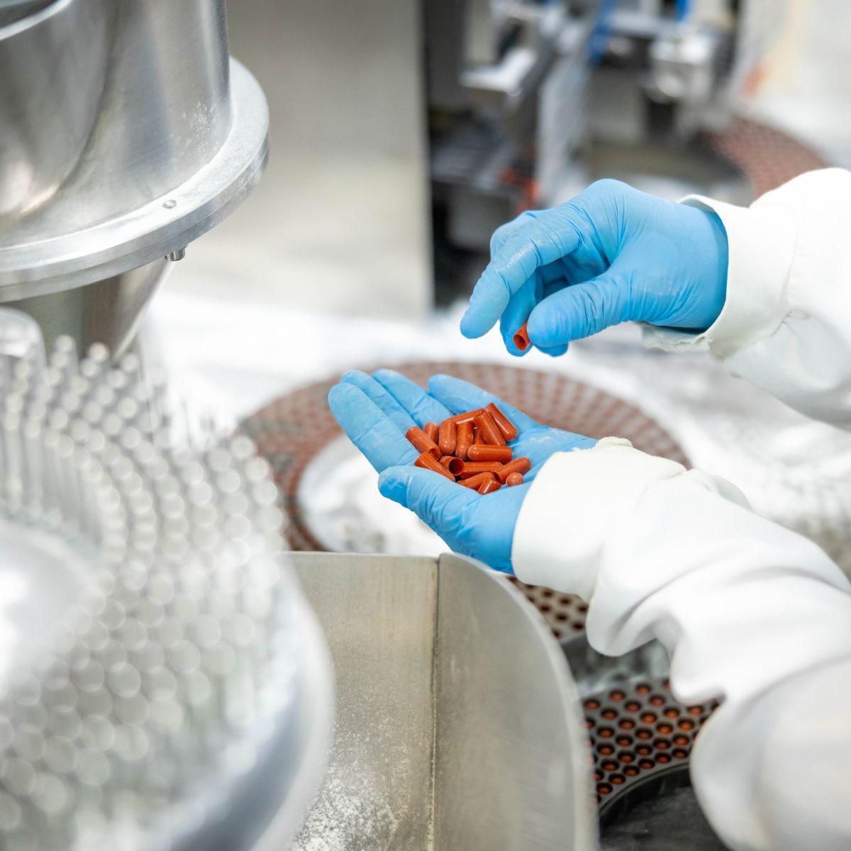 A technician wearing blue gloves inspects orange pharmaceutical capsules near a filling machine in a sterile manufacturing facility.