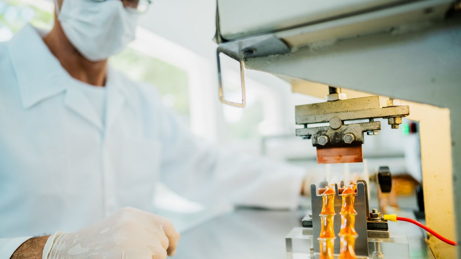 A technician in protective gear monitors an automated machine processing amber vials in a sterile laboratory environment.