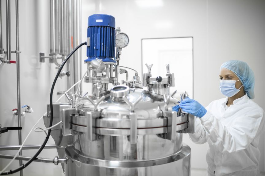 A technician in cleanroom attire operates a stainless steel industrial mixing vessel in a sterile pharmaceutical manufacturing facility.