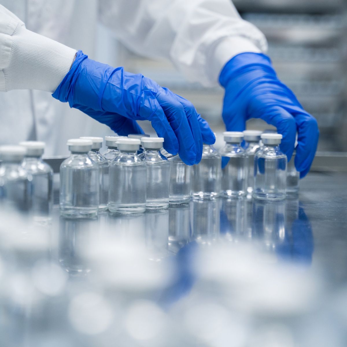 A lab technician in protective gear inspects clear glass medical vials on a stainless steel conveyor line in a sterile pharmaceutical manufacturing facility.
