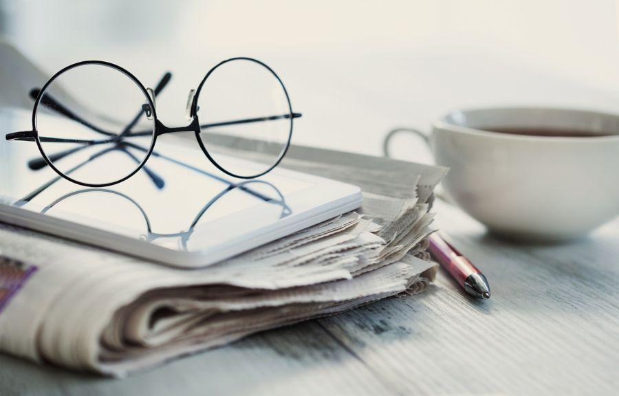 Close-up of glasses resting on a tablet, surrounded by a newspaper, pen, and a cup of coffee.