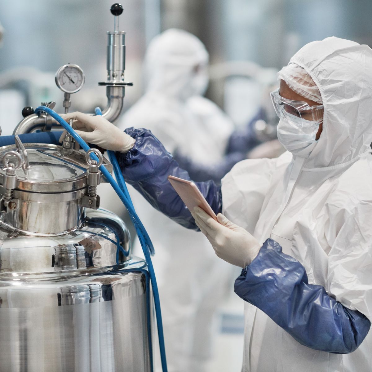 A lab technician in protective clothing checks equipment while holding a tablet in a high-tech facility.