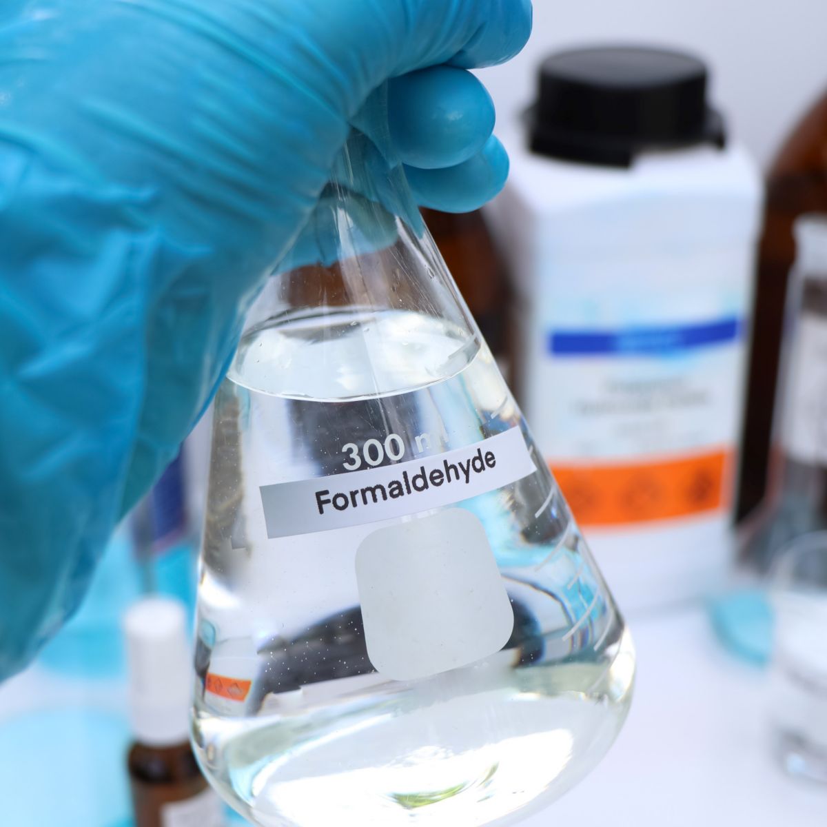 A lab technician in blue gloves holds a labeled flask of formaldehyde, surrounded by various lab materials.