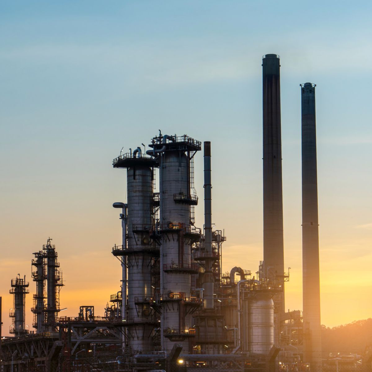  A silhouetted image of an industrial facility at sunset, featuring multiple towers and smoke stacks.