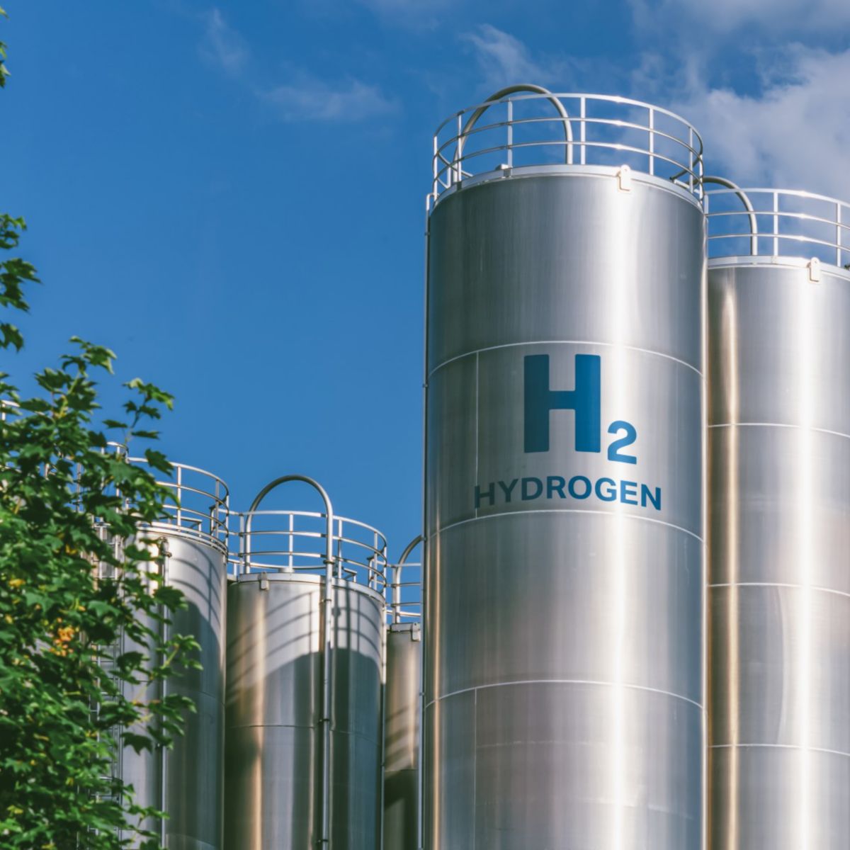 A view of tall hydrogen storage tanks marked with "H2," framed by a bright sky and greenery.
