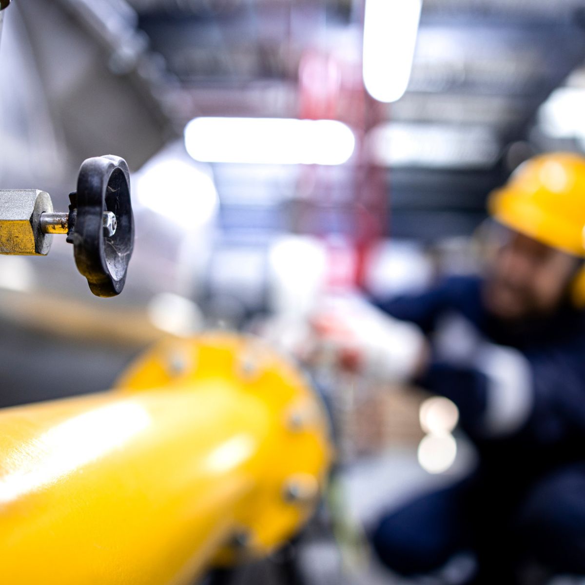 Detailed shot of a valve on a yellow pipe, with an industrial worker visible in the background.