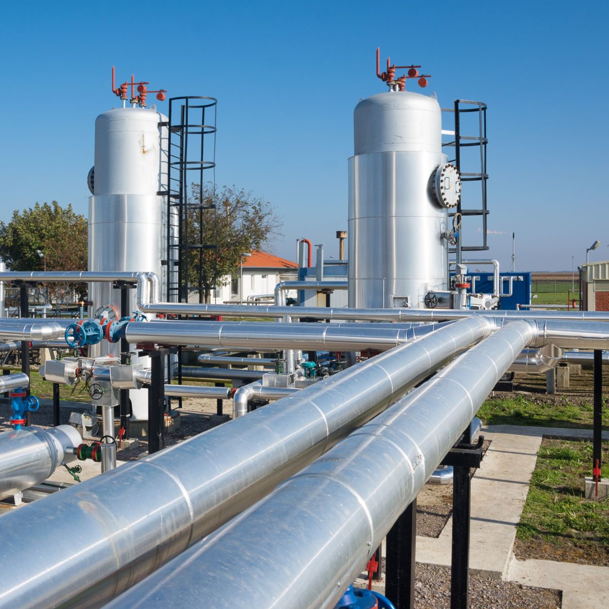 A gas processing facility featuring shiny pipelines, large storage tanks, and a clear blue sky in the background.