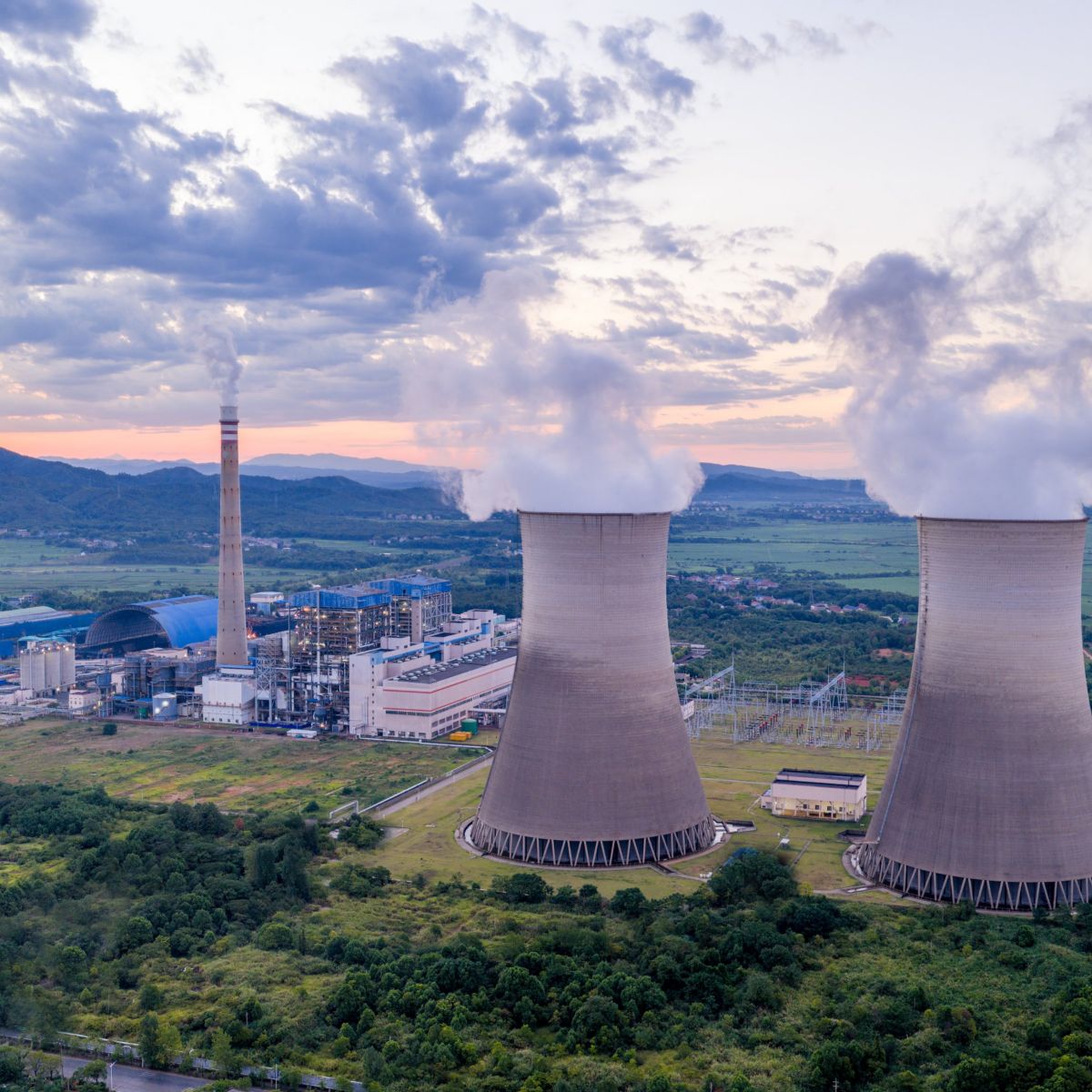 An aerial view captures a thermal power plant at sunset, highlighting its massive cooling towers emitting steam amidst a rural landscape.