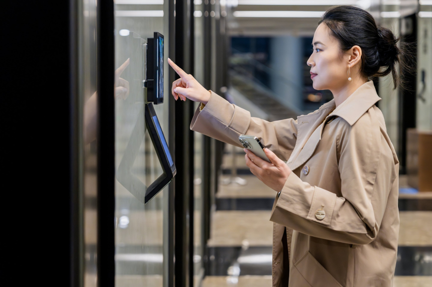 Business woman in a trench coat uses a biometric digital interface to access a secured office space while holding her smartphone.