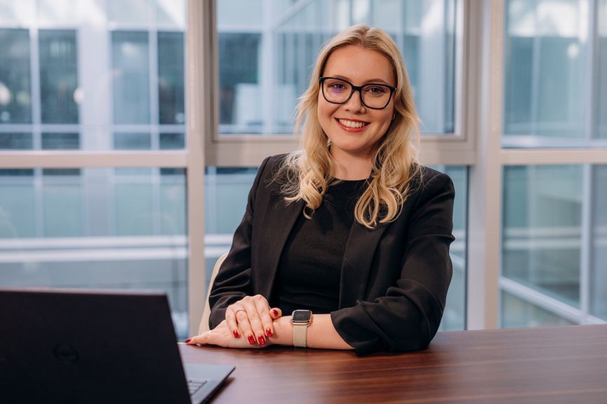 A smiling professional woman in a suit poses at her desk in a modern, light-filled office environment.
