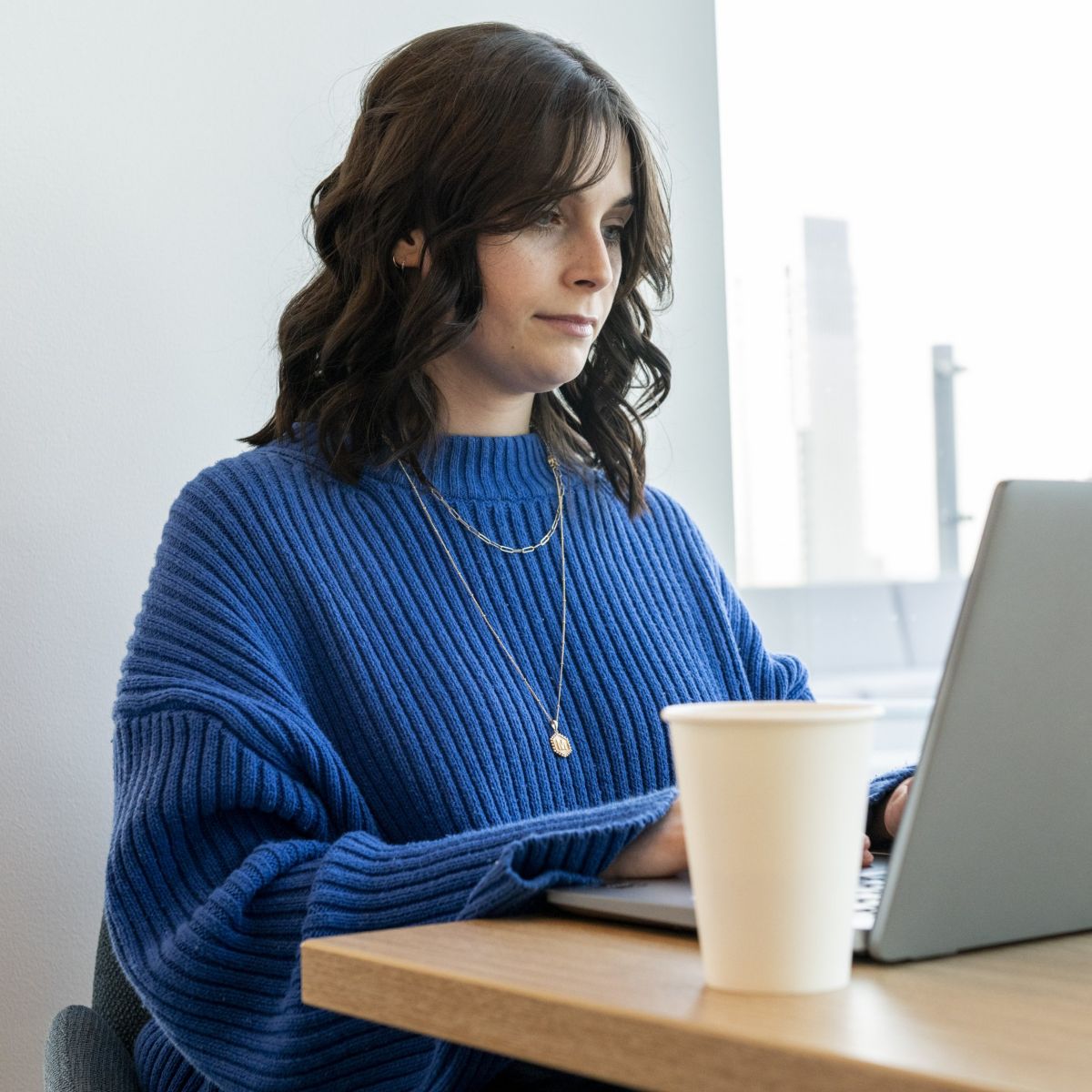 A young woman in a blue sweater works on her laptop at a wooden desk with a coffee cup.