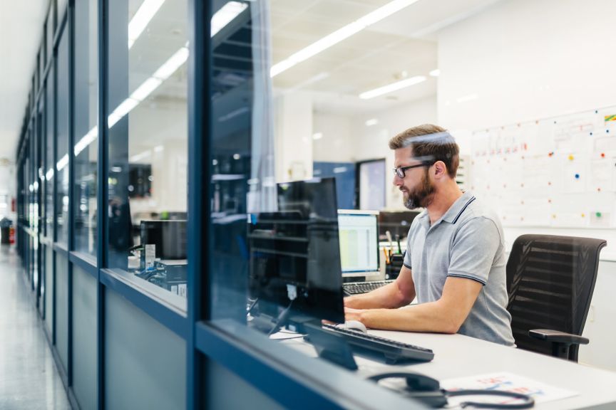 A focused male employee in a modern office uses a computer while seen through a glass partition.