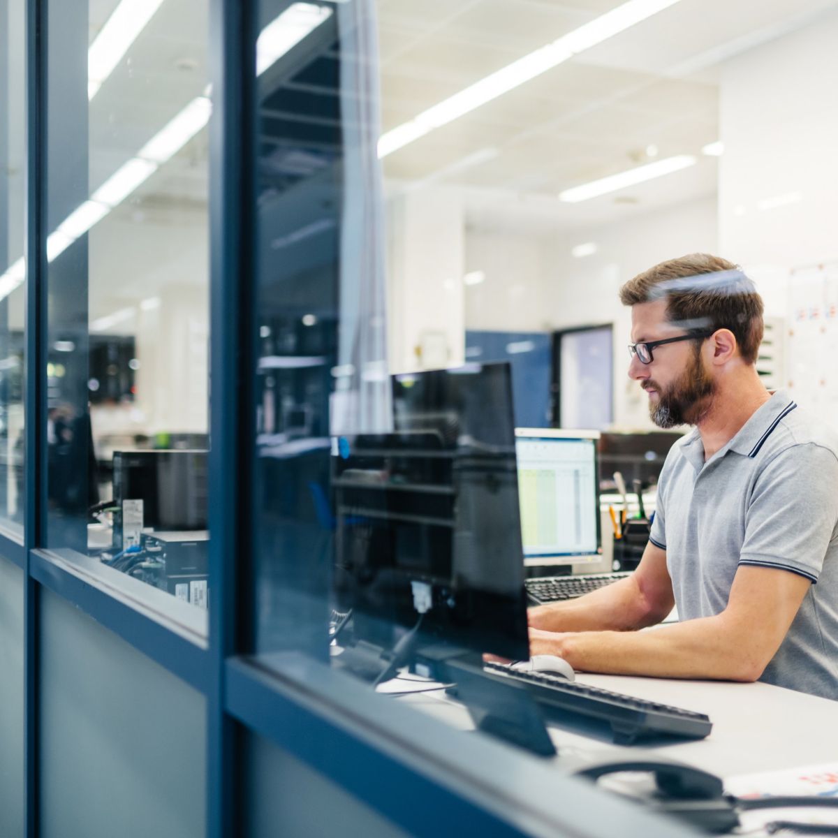 A focused male employee in a modern office uses a computer while seen through a glass partition.