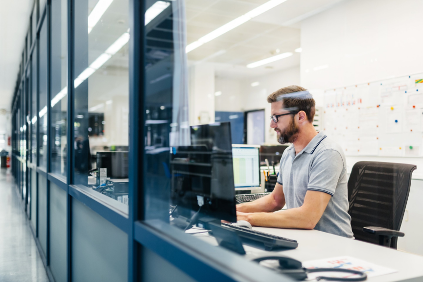 A focused male employee in a modern office uses a computer while seen through a glass partition.