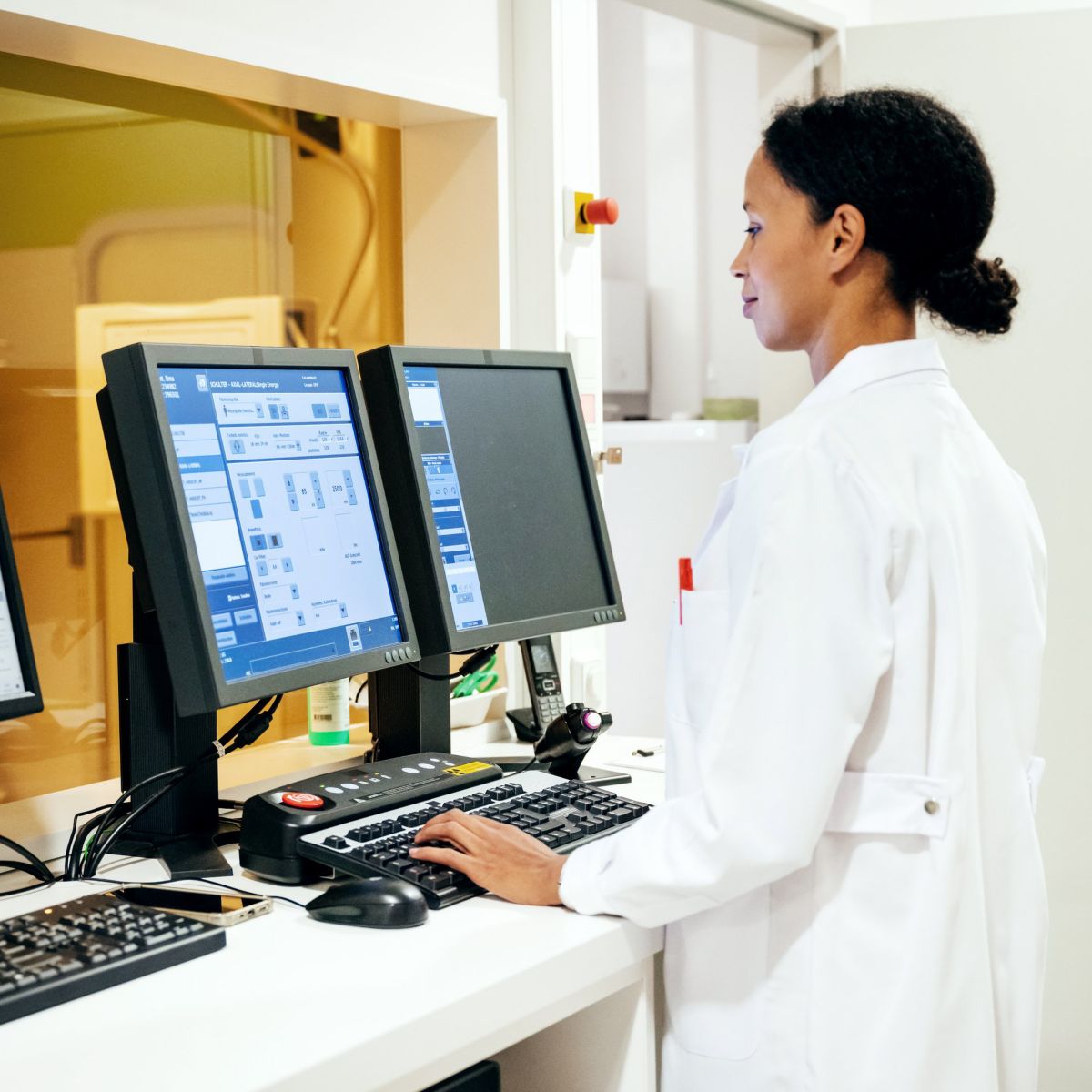 A professional radiologist in a white lab coat monitors medical imaging data at a computer workstation in a clinical diagnostic environment.