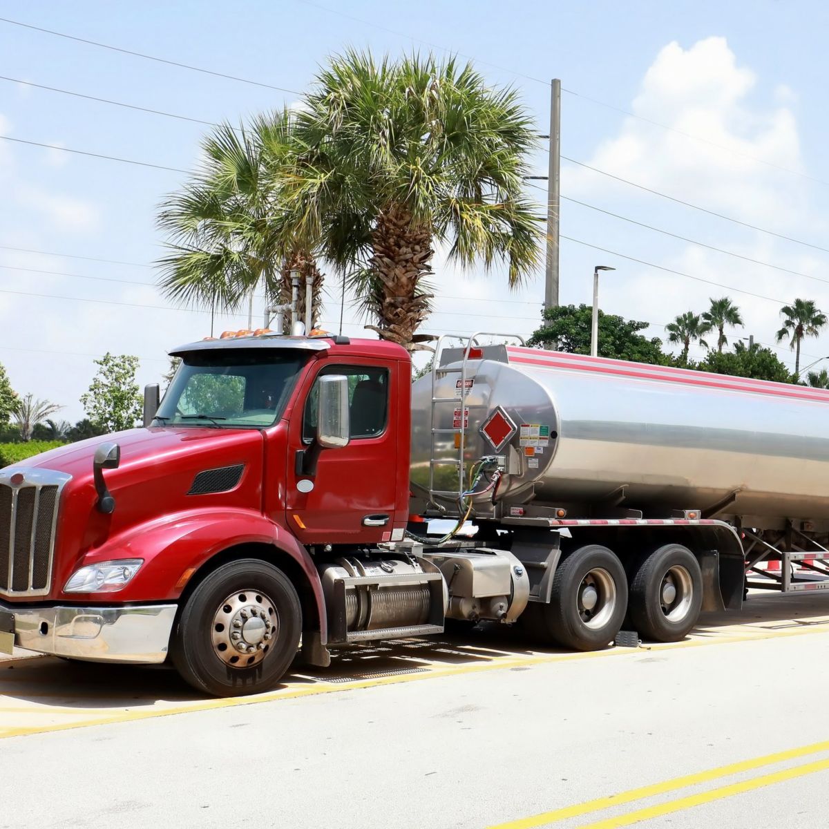 A bright red industrial fuel tanker truck is parked on an asphalt lot near palm trees on a clear, sunny day.