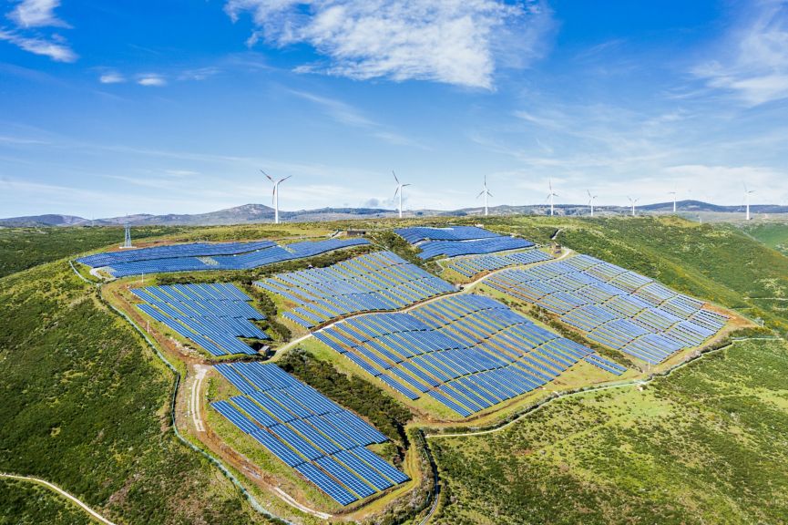 Aerial view of a solar farm integrated with wind turbines on a scenic green hillside.