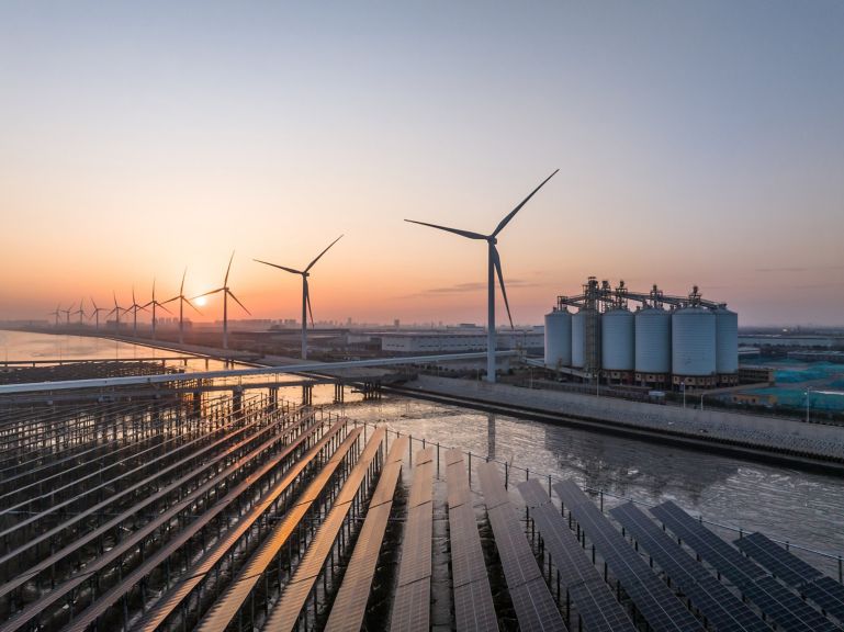 Solar panels and wind turbines capture energy alongside industrial storage silos at sunrise.