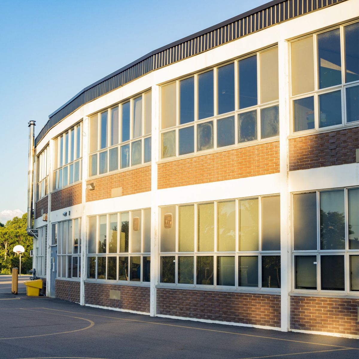 A brick school exterior features large windows and multiple basketball hoops on an expansive asphalt playground during golden hour.