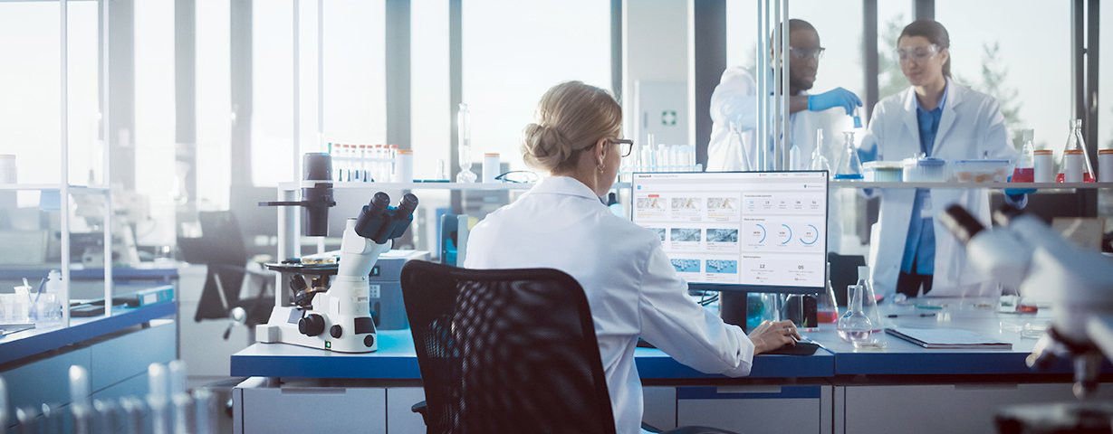 A seated scientist reviews multiple screens at a laboratory workstation while colleagues work nearby.