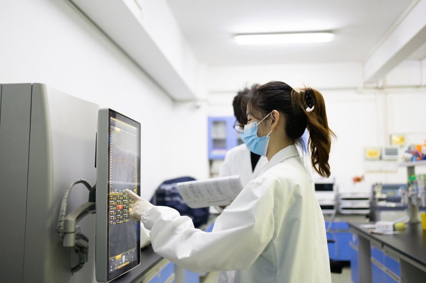 Two researchers in lab coats and masks operate an analytical instrument in a clean, professional research facility.