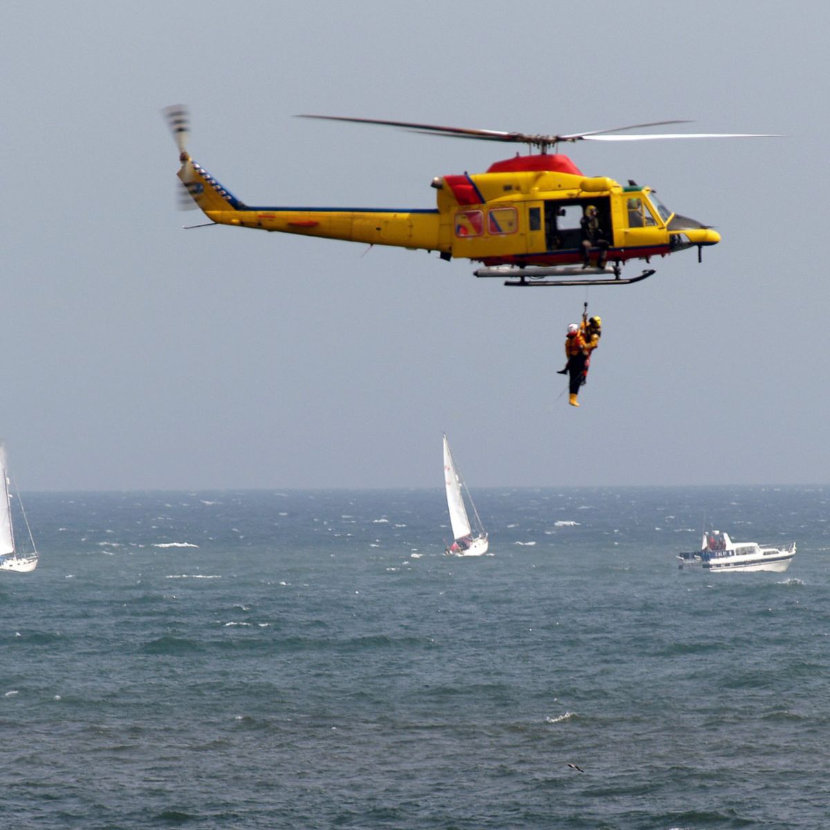 A search and rescue helicopter performs a hoist operation over the sea with sailboats in the background.