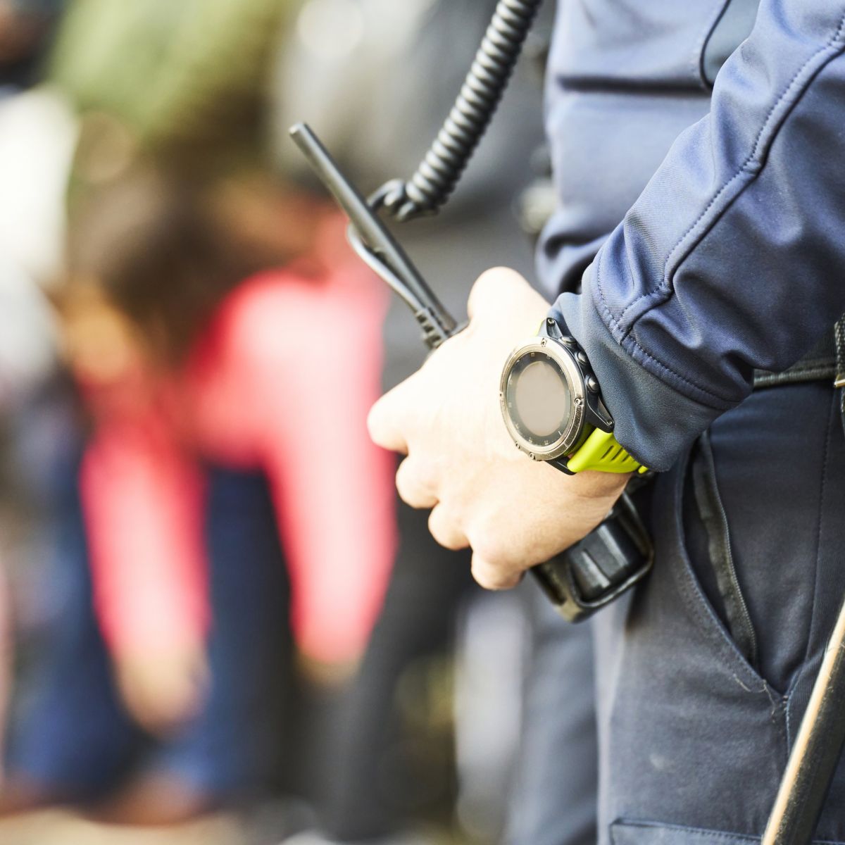 A security officer stands ready with a two-way radio and tactical watch while monitoring a crowd of people in the blurred background.