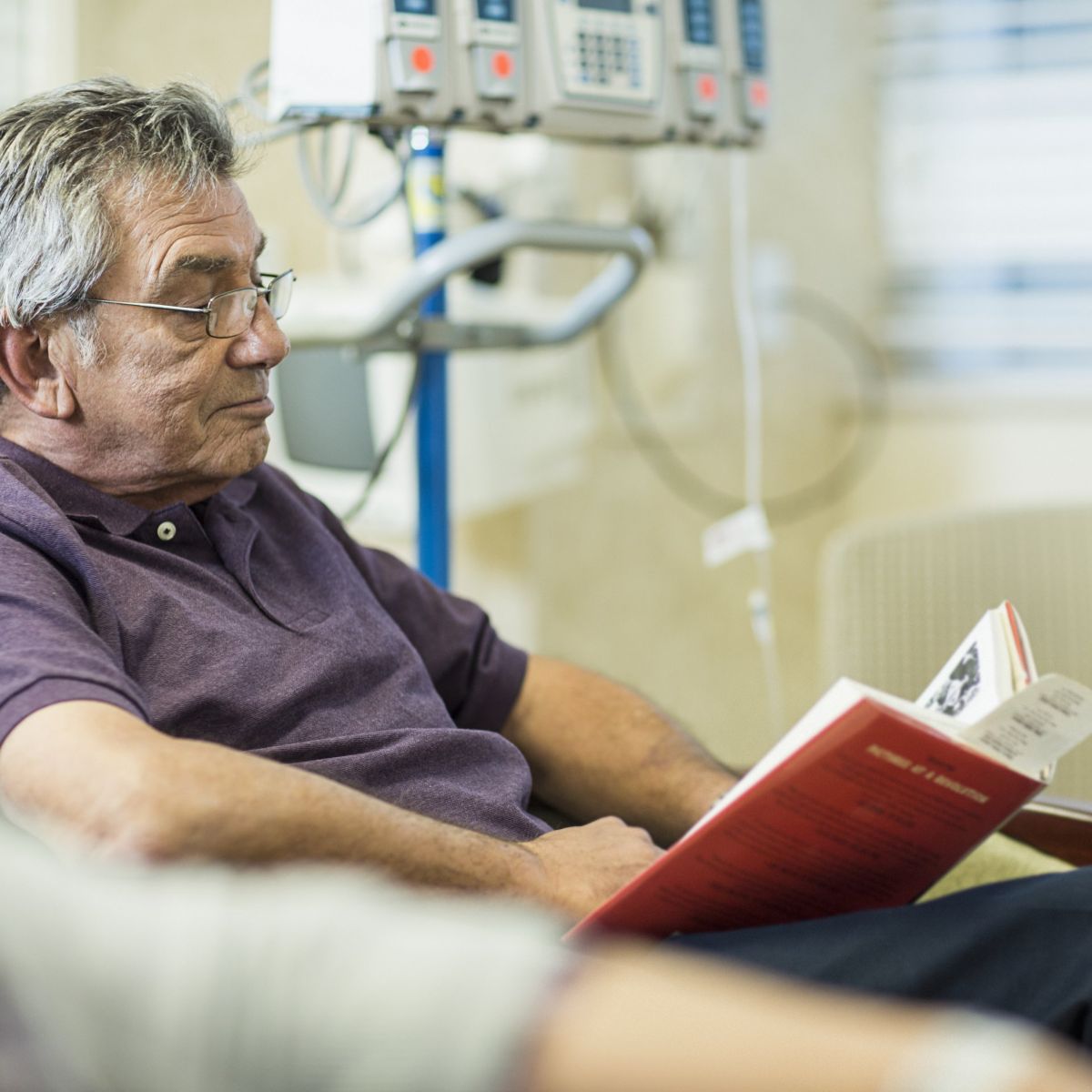 An elderly man in a clinical setting sits in a chair reading a book while receiving medical treatment.