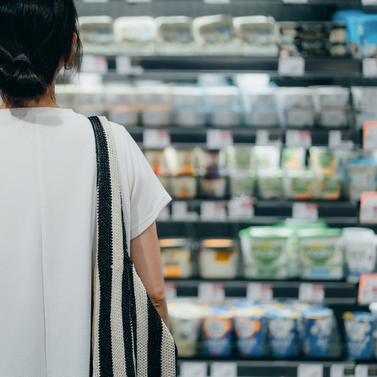 A woman with a shoulder bag stands before a well-stocked grocery store refrigerator shelf, carefully selecting dairy products.