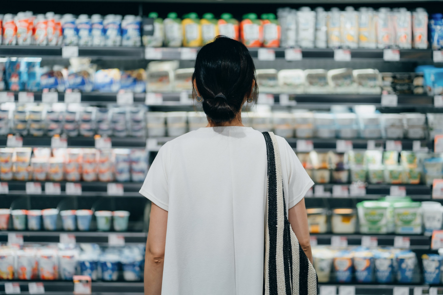 A woman with a shoulder bag stands before a well-stocked grocery store refrigerator shelf, carefully selecting dairy products.