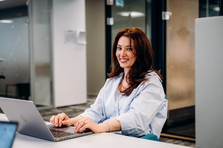 A professional woman in a light blue shirt smiles while typing on a laptop at her desk in a modern corporate office environment.