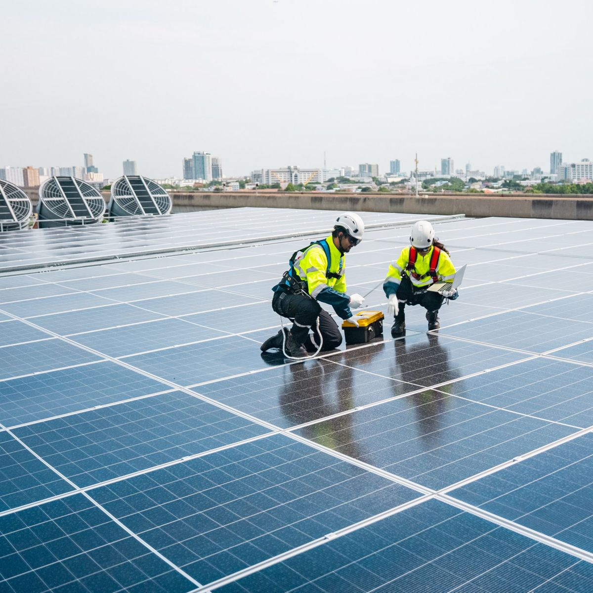 Two technicians in safety gear inspect solar panels on a building rooftop against a city skyline.